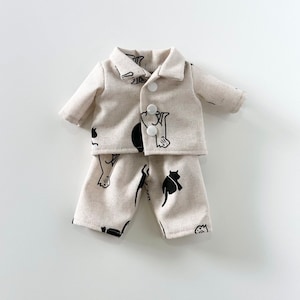Puede incluir: Un conjunto de pijama beige con ilustraciones de gatos negros. La parte superior tiene un cierre de bot&oacute;n y los pantalones tienen un ajuste holgado.