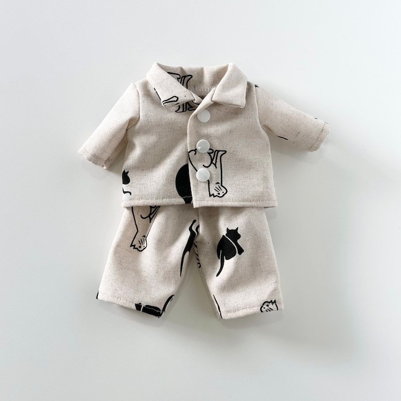 Puede incluir: Un conjunto de pijama beige con ilustraciones de gatos negros. La parte superior tiene un cierre de bot&oacute;n y los pantalones tienen un ajuste holgado.