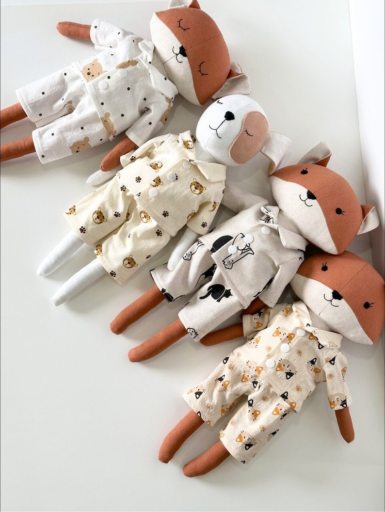 Puede incluir: Cuatro juguetes de peluche de zorro con pijamas con diferentes estampados de animales. Las pijamas son beige con estampados de animales negros y marrones. Los zorros son marrones con detalles blancos y los ojos cerrados.