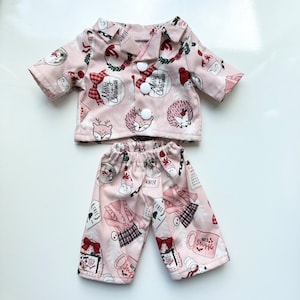 Puede incluir: Un conjunto de pijama rosa con una camisa de manga larga y pantalones. La camisa tiene un frente abotonado y presenta un estampado navide&ntilde;o festivo con acentos rojos y blancos. Los pantalones tienen un estampado similar con una variedad de ilustraciones de tem&aacute;tica invernal.