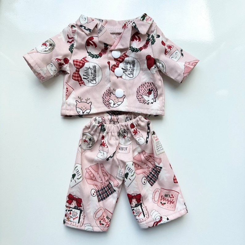 Puede incluir: Un conjunto de pijama rosa con una camisa de manga larga y pantalones. La camisa tiene un frente abotonado y presenta un estampado navide&ntilde;o festivo con acentos rojos y blancos. Los pantalones tienen un estampado similar con una variedad de ilustraciones de tem&aacute;tica invernal.