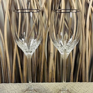 May include: Two clear wine glasses with silver rims. The glasses have a detailed, textured design on the bowl and a long, slender stem. They are set against a backdrop of brown, natural-looking branches.