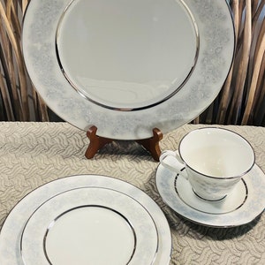 May include: A set of white porcelain dinnerware with a delicate silver trim and a floral pattern. The set includes a large dinner plate, a smaller salad plate, and a teacup and saucer.