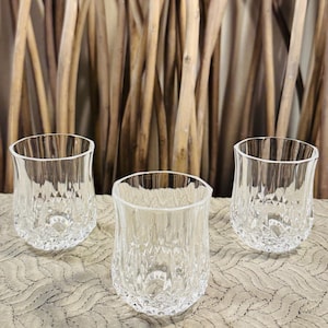 May include: Three clear, textured drinking glasses with a diamond pattern. The glasses have a slightly curved shape and sit on a patterned surface. The background features vertical wooden sticks.