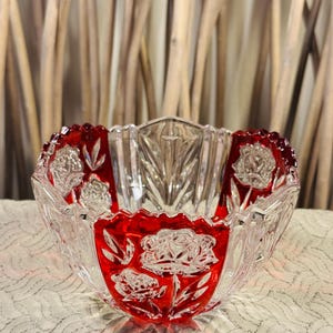May include: A clear and ruby red glass bowl featuring a scalloped rim and intricate relief carvings of rose blossoms and leaves. The bowl has alternating panels of transparent glass and deep red glass, showcasing a detailed floral pattern.