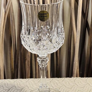 May include: A clear, stemmed wine glass with a textured, diamond-cut design on the bowl. The glass has a round base and a slender stem. A gold label with the text "Cristal d'Arques" is affixed to the bowl.