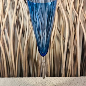 May include: A tall, elegant champagne flute with a vibrant blue bowl and a clear, slender stem. The glass features vertical, textured lines within the blue bowl. The base is a clear, circular foot. The background is a woven, light brown pattern.