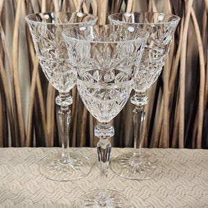 May include: Three clear crystal wine glasses featuring intricate cut glass patterns with starburst and diamond motifs. Each glass has a tall, slender stem and a wide, circular base with a matching starburst design etched into the bottom surface.