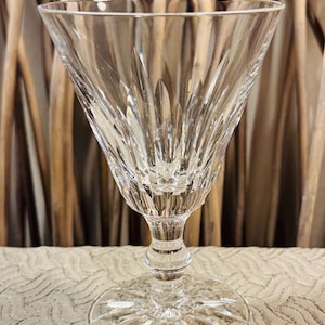 May include: A clear, cut-crystal wine glass with a wide, flared bowl and a detailed, patterned stem and base. The glass features intricate, radiating cuts that catch the light, creating a sparkling effect. The background is a neutral beige.