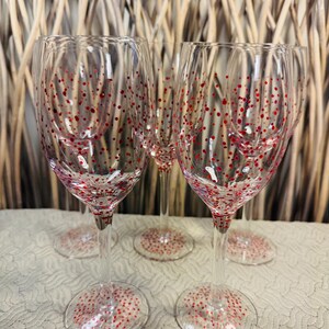 May include: Clear wine glasses with red polka dots. The glasses have a long stem and a round base, also decorated with red dots. The background features a woven, light brown texture.