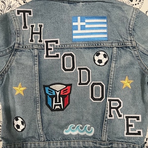 May include: A blue denim jacket with various patches, including the name "Theodore", a Greek flag, a soccer ball, a robot, a wave, and two gold stars.