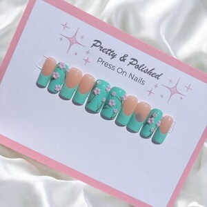 Könnte beinhalten: Ein Set von Press-On-Nägeln mit helltürkisfarbener Basis und einem nudefarbenen Tipp. Die Nägel sind mit kleinen rosa Blüten verziert. Der Text "Pretty & Polished Press On Nails" ist auf der Karte zu sehen.