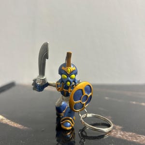 May include: A miniature warrior figurine in blue and gold, holding a silver sword and a shield. The figurine is attached to a silver keyring. The warrior has a helmet and yellow eyes.