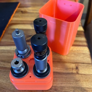 May include: An orange plastic case with four metal reloading dies. The case has a lid that is also orange. The dies are different sizes and have black and silver accents.