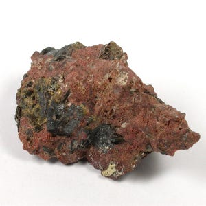 May include: A raw mineral specimen featuring a porous, reddish-brown rock matrix embedded with clusters of dark, metallic, needle-like crystals. The stone has an irregular, jagged shape with visible crystalline formations scattered across its surface.