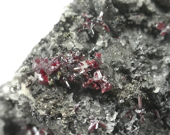 Könnte beinhalten: Ein Mineralexemplar mit Ansammlungen tiefroter, prismatischer Kristalle, die in eine dunkle, texturierte, anthrazitfarbene Matrix eingebettet sind. Die Oberfläche zeigt eine Mischung aus metallischen, kristallinen und körnigen Texturen mit kleinen, reflektierenden, dunklen Mineraleinschlüssen.