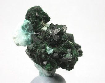Könnte beinhalten: Ein Mineralexemplar mit einer Ansammlung von dunkelwaldgrünen, kristallinen Strukturen mit scharfen, kantigen Rändern. Die Kristalle sind dicht gepackt, weisen einen metallischen Glanz auf und sind auf einer helleren, mintgrünen Basis befestigt.