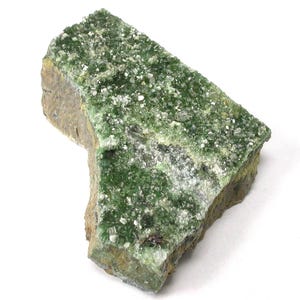 Pode incluir: Um espécime mineral natural em bruto. A pedra apresenta uma base de rocha castanha e cinzenta, com uma vibrante e brilhante formação de cristais verdes que cobre a superfície. Pequenas inclusões cristalinas brancas estão espalhadas.