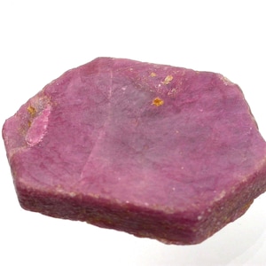 May include: A raw, hexagonal-shaped ruby crystal with a deep magenta-pink color. The stone features a flat, textured surface with visible natural inclusions and a slightly irregular, rough-hewn edge, showcasing its unpolished, natural state.