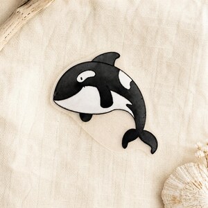 Könnte beinhalten: Ein gestanzter Aufkleber mit einer schwarz-weißen Illustration eines Orcas. Der Orca ist im Seitenprofil mit gekrümmtem Körper, einer markanten Rückenflosse und einem weißen Augenfleck dargestellt, ausgeführt in einer Aquarell-Optik.