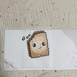 May include: A whimsical illustration of a smiling, cartoon-style cracker with large eyes and a bite taken out. The cracker is light brown with darker brown edges and small dots. The background is white with a light beige fabric.