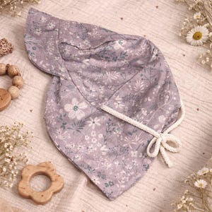 Peut inclure: Un bonnet de bébé lavande avec un motif floral de fleurs et de feuilles blanches et vertes. Le bonnet a un lien blanc et est fait de tissu doux. Un jouet en bois en forme de fleur et un bracelet de perles en bois sont également dans l'image.