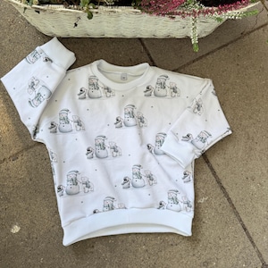 Könnte beinhalten: Weißes Sweatshirt mit einem sich wiederholenden Muster aus Schneemännern, Pinguinen und Sternen. Das langärmlige Oberteil hat einen Rundhalsausschnitt und gerippte Bündchen. Das Design zeigt Schneemänner mit Hüten und Schals.