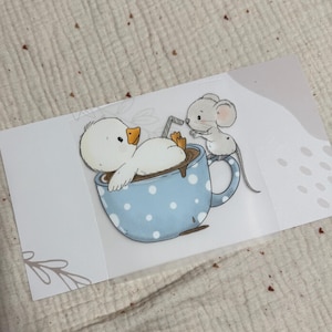 May include: A charming illustration depicts a white duck lounging in a blue polka-dot mug filled with a brown liquid. A small grey mouse is positioned on the mug's rim, holding a straw. The artwork is on a white card with a floral design.