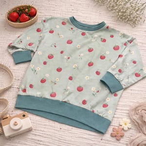 Könnte beinhalten: Hellgrünes Sweatshirt mit einem Muster aus roten Erdbeeren und weißen Blüten. Die Bündchen, der Halsausschnitt und der Saum sind tealfarben. Die Ärmel sind dreiviertellang.