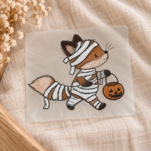 Könnte beinhalten: Ein quadratischer Aufkleber mit einem Cartoon-Fuchs, der als Mumie in weiße Bandagen gewickelt ist und einen kleinen orangefarbenen Kürbis-Eimer hält. Der Fuchs hat braunes Fell, dunkle Ohren und einen buschigen Schwanz, alles in Stoffstreifen.