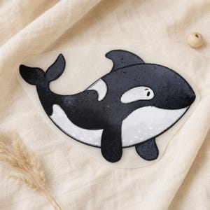 Könnte beinhalten: Ein flacher, gestanzter Aufkleber oder Ausschnitt mit einer stilisierten Illustration eines Orcas. Der Orca ist in Schwarz-Weiß gehalten und zeigt einen abgerundeten Körper, eine Rückenflosse und ein kleines Auge in einem klaren Grafikstil.