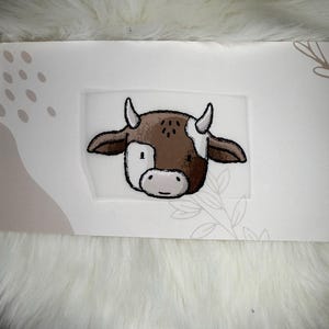 Könnte beinhalten: Ein Aufkleber mit dem Kopf einer Cartoon-Kuh. Die Kuh hat braune und weiße Markierungen, schwarze Umrisse und kleine weiße Hörner. Der Aufkleber befindet sich auf einem weißen Hintergrund mit einem dekorativen Rand.