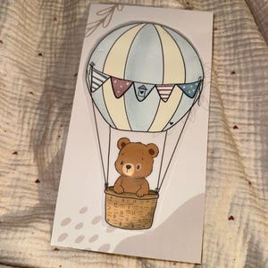 May include: A whimsical illustration of a brown teddy bear in a wicker basket suspended from a pastel blue and cream hot air balloon. The balloon is decorated with colorful pennants. The image is on a white card.