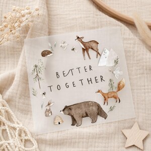 Könnte beinhalten: Ein quadratisches, durchscheinendes Schild mit dem Text BETTER TOGETHER in schwarzer Schrift. Das Design enthält Aquarellillustrationen eines Rehs, eines Fuchses, eines Bären, eines Hasen, eines Igels, eines Bienenstocks, Bäumen und Bergen.