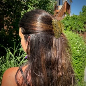 May include: A translucent olive green hair claw clip, shaped like a leaf, secures a section of long, brown hair. The clip, made of shiny plastic, is positioned on the right side of the head. The hair has highlights.