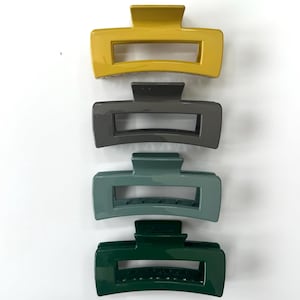 May include: Four hair clips in different colors: mustard yellow, gray, teal, and forest green. The clips are all the same shape and size, with a rectangular base and a curved top.