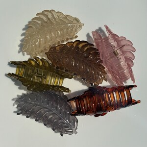 May include: Six translucent leaf-shaped hair claw clips in various colours: light tan, brown, pink, olive green, dark brown, and grey. Each clip has a detailed leaf vein design and a spring-loaded clasp for securing hair.