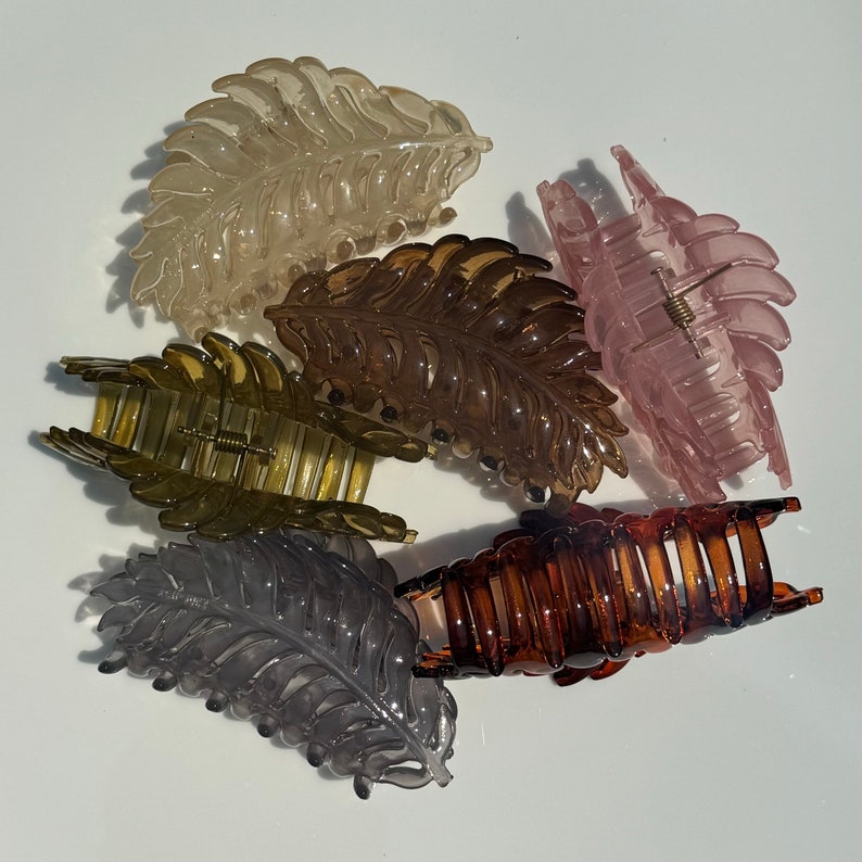 May include: Six translucent leaf-shaped hair claw clips in various colours: light tan, brown, pink, olive green, dark brown, and grey. Each clip has a detailed leaf vein design and a spring-loaded clasp for securing hair.