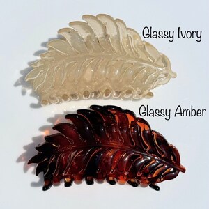 May include: Two leaf-shaped hair clips are shown. One is a translucent ivory colour, and the other is a glossy amber. The clips are made of a shiny material and are designed to resemble a leaf. The text "Glassy Ivory" and "Glassy Amber" are written on the image.