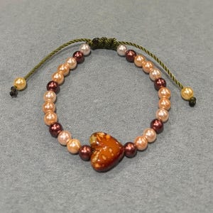 May include: A beaded bracelet featuring a heart-shaped charm. The bracelet is made with peach, burgundy, and white beads. The heart charm is a warm brown color with a glossy finish. The bracelet has an adjustable green cord.
