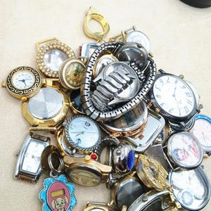 K&ouml;nnte beinhalten: Eine Sammlung verschiedener Armbanduhren und Uhrenteile, darunter diverse runde und rechteckige Geh&auml;use, Metallgliederarmb&auml;nder und Zifferbl&auml;tter mit analogen Anzeigen, teilweise mit dekorativen gold- und silberfarbenen Akzenten.