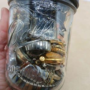 K&ouml;nnte beinhalten: Ein klares Ball Wide Mouth Einmachglas aus Glas mit schwarzem Deckel, gef&uuml;llt mit einer Sammlung verschiedener Armbanduhren und Uhrenarmb&auml;ndern aus Metall in Silber- und Goldt&ouml;nen, mit verschiedenen Zifferbl&auml;ttern und Gliederdesigns.