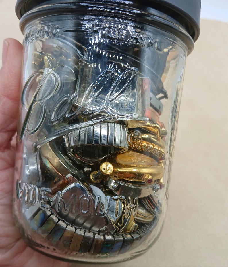 K&ouml;nnte beinhalten: Ein klares Ball Wide Mouth Einmachglas aus Glas mit schwarzem Deckel, gef&uuml;llt mit einer Sammlung verschiedener Armbanduhren und Uhrenarmb&auml;ndern aus Metall in Silber- und Goldt&ouml;nen, mit verschiedenen Zifferbl&auml;ttern und Gliederdesigns.