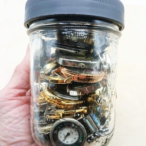 K&ouml;nnte beinhalten: Ein durchsichtiges Einmachglas aus Glas mit einem dunkelgrauen, gerippten Schraubdeckel, gef&uuml;llt mit einer Sammlung verschiedener Metall-Uhrengeh&auml;use, Uhrenteilen und einer sichtbaren runden Armbanduhr mit wei&szlig;em Zifferblatt am Boden.