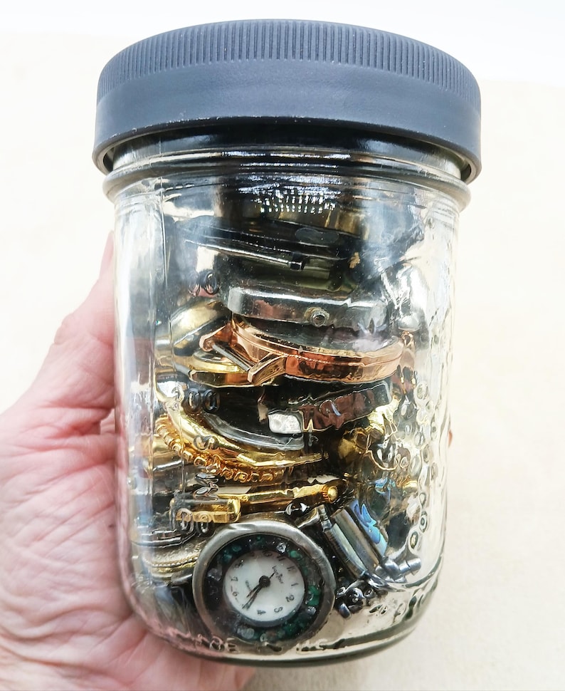 K&ouml;nnte beinhalten: Ein durchsichtiges Einmachglas aus Glas mit einem dunkelgrauen, gerippten Schraubdeckel, gef&uuml;llt mit einer Sammlung verschiedener Metall-Uhrengeh&auml;use, Uhrenteilen und einer sichtbaren runden Armbanduhr mit wei&szlig;em Zifferblatt am Boden.