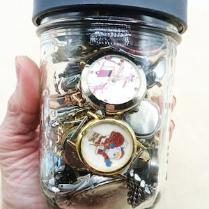 K&ouml;nnte beinhalten: Ein durchsichtiges Einmachglas aus Glas mit schwarzem Deckel, gef&uuml;llt mit verschiedenen Armbanduhren. Zwei Uhren sind deutlich zu sehen: eine mit wei&szlig;em Zifferblatt und rosa Grafik sowie eine mit goldfarbenem Geh&auml;use und Weihnachtsmann-Motiv.
