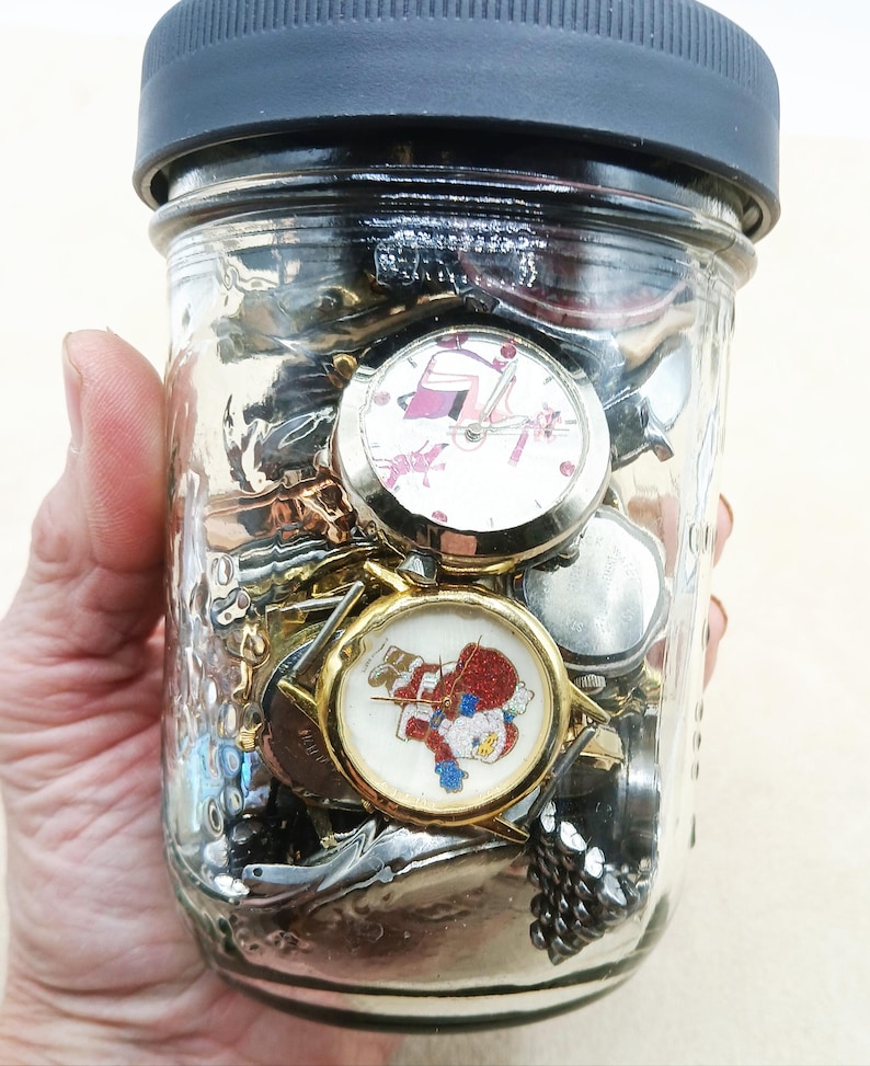 K&ouml;nnte beinhalten: Ein durchsichtiges Einmachglas aus Glas mit schwarzem Deckel, gef&uuml;llt mit verschiedenen Armbanduhren. Zwei Uhren sind deutlich zu sehen: eine mit wei&szlig;em Zifferblatt und rosa Grafik sowie eine mit goldfarbenem Geh&auml;use und Weihnachtsmann-Motiv.