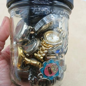 K&ouml;nnte beinhalten: Ein klares Glasgef&auml;&szlig; mit schwarzem Deckel, gef&uuml;llt mit einer Sammlung verschiedener Armbanduhren aus Metall und einem kleinen blau-rosa Emaille-Anh&auml;nger mit einem Cartoon-Schwein. Das Glas enth&auml;lt mehrere Zifferbl&auml;tter, Metallb&auml;nder und Geh&auml;use.