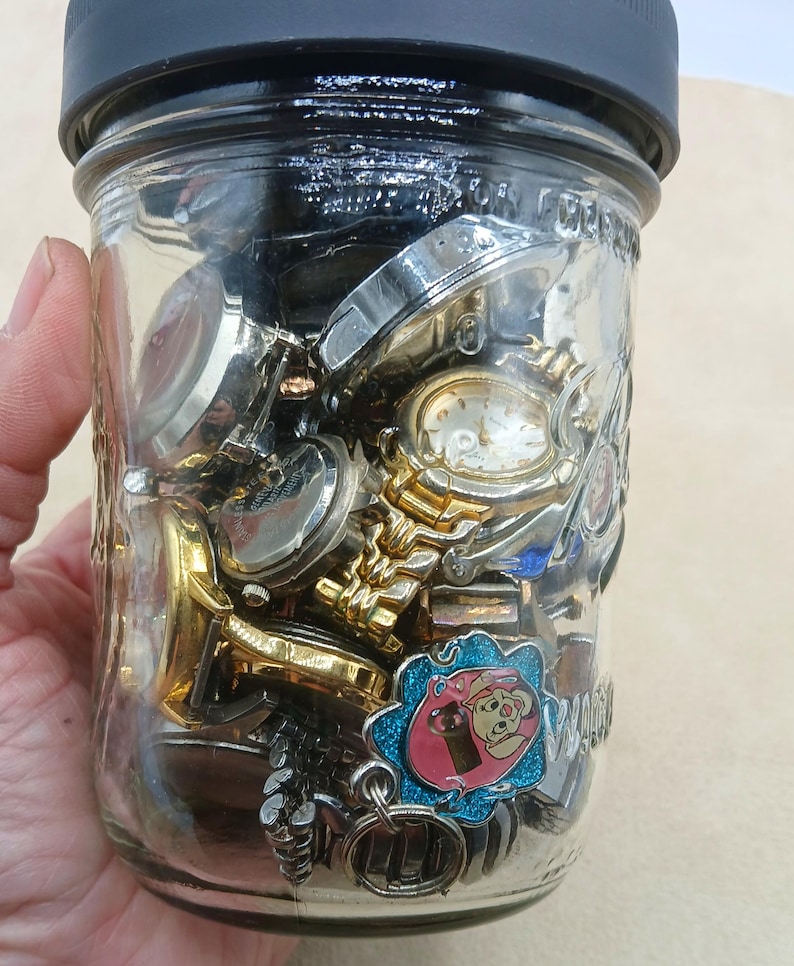 K&ouml;nnte beinhalten: Ein klares Glasgef&auml;&szlig; mit schwarzem Deckel, gef&uuml;llt mit einer Sammlung verschiedener Armbanduhren aus Metall und einem kleinen blau-rosa Emaille-Anh&auml;nger mit einem Cartoon-Schwein. Das Glas enth&auml;lt mehrere Zifferbl&auml;tter, Metallb&auml;nder und Geh&auml;use.