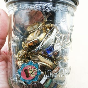 K&ouml;nnte beinhalten: Ein durchsichtiges Ball-Einmachglas aus Glas mit einem dunkelgrauen Deckel, gef&uuml;llt mit verschiedenen Metallgegenst&auml;nden, darunter goldfarbene Uhrenteile und ein blau glitzernder Anh&auml;nger mit einer rosa Zeichentrickfigur.
